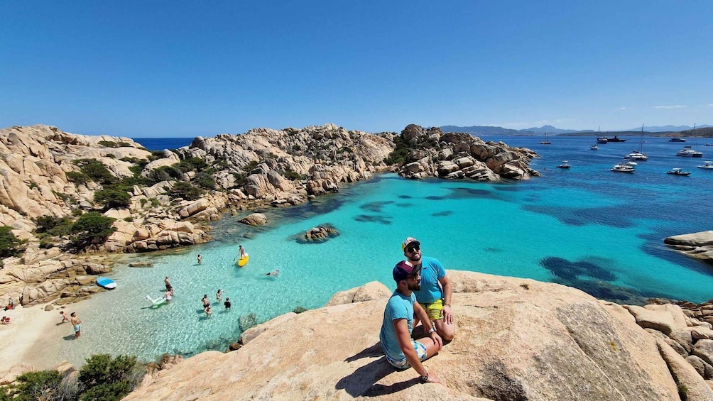 A rocky coastline with clear turquoise waters, people swimming and sunbathing, and boats anchored offshore. 2