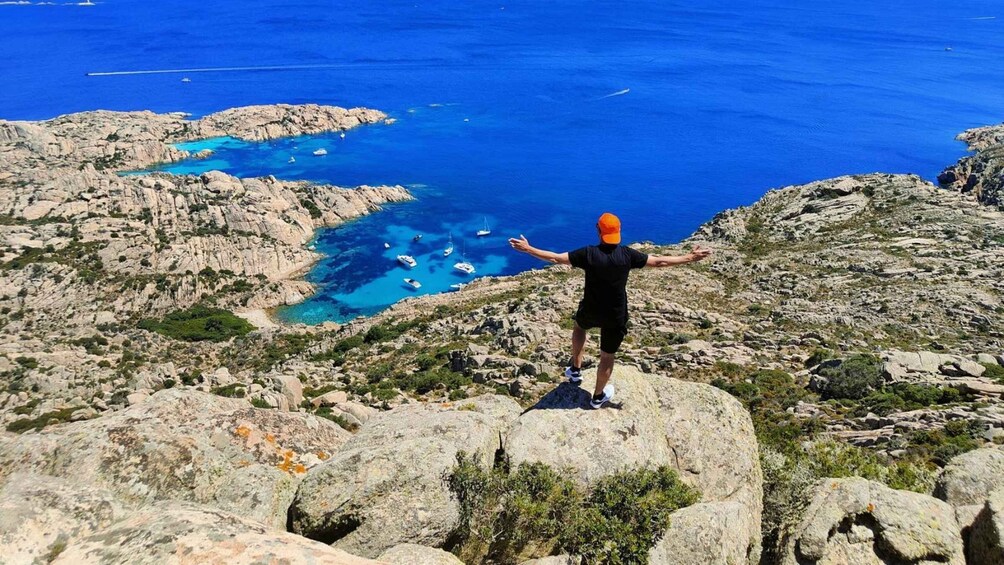 A person standing on a rocky outcrop with arms outstretched, overlooking a coastal area with clear blue waters and boats. 1