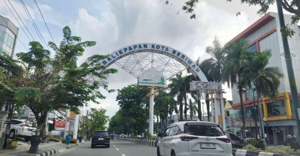 A street with a large archway labeled 'BALI KOTA BERHAN' and cars parked on the side. 1