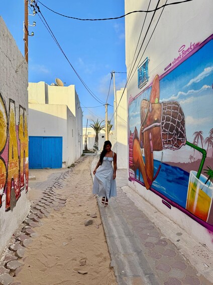 A narrow alley with a mural of a beach scene, a blue gate, and a person walking. 4