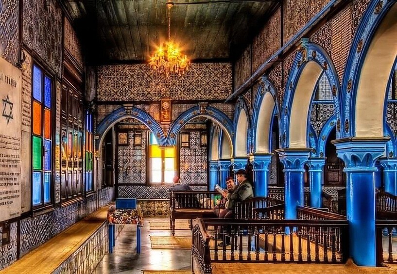 An ornate synagogue interior with blue arches, a chandelier, and Hebrew inscriptions. 1