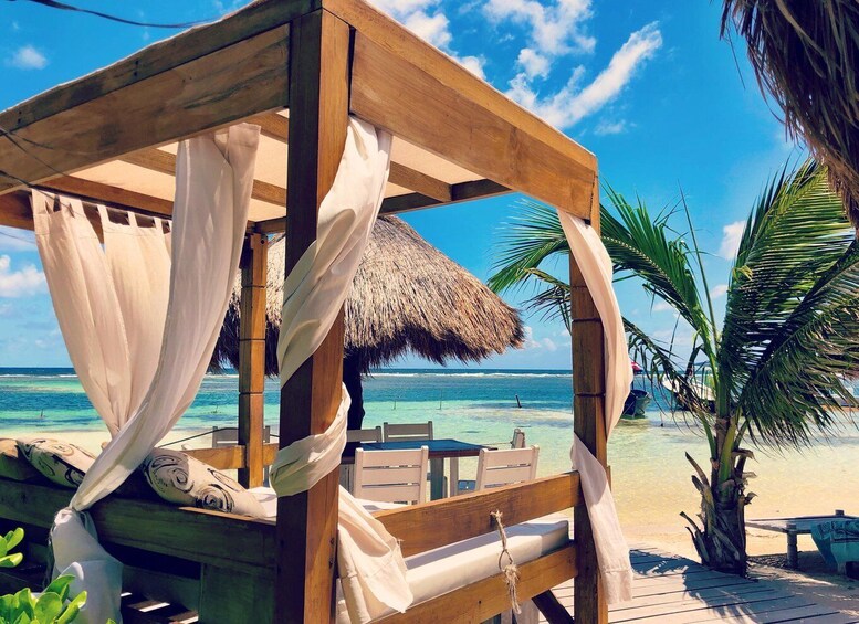 A wooden beach cabana with a thatched roof, white curtains, and a bench. 1