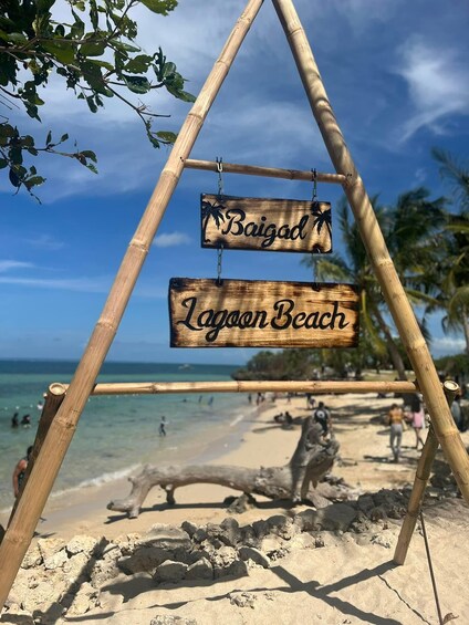 A wooden sign with 'Baigad' and 'Lagoon Beach' written on it, set against a beach backdrop with palm trees and the ocean. 3