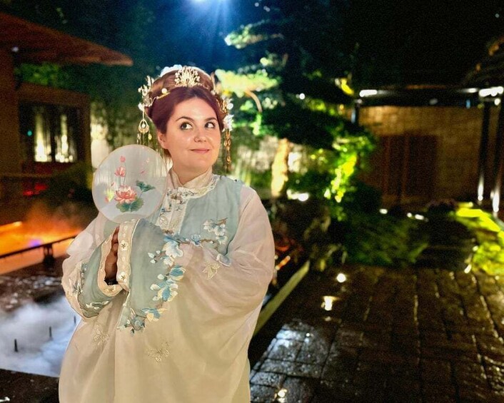 A person in a traditional dress holding a fan, standing on a cobblestone path with greenery and a building in the background. 1