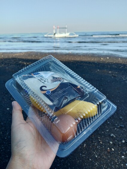 A hand holding a sandwich in a plastic container with a beach and ocean in the background. 3