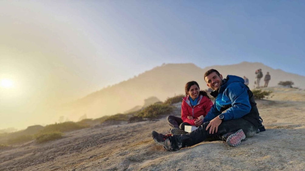 Two people sitting on a rocky surface with a mountain in the background. 4