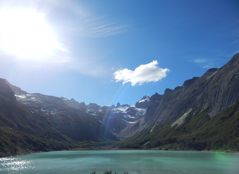A mountain lake with clear turquoise water, surrounded by steep, forested mountains under a bright sun. 1