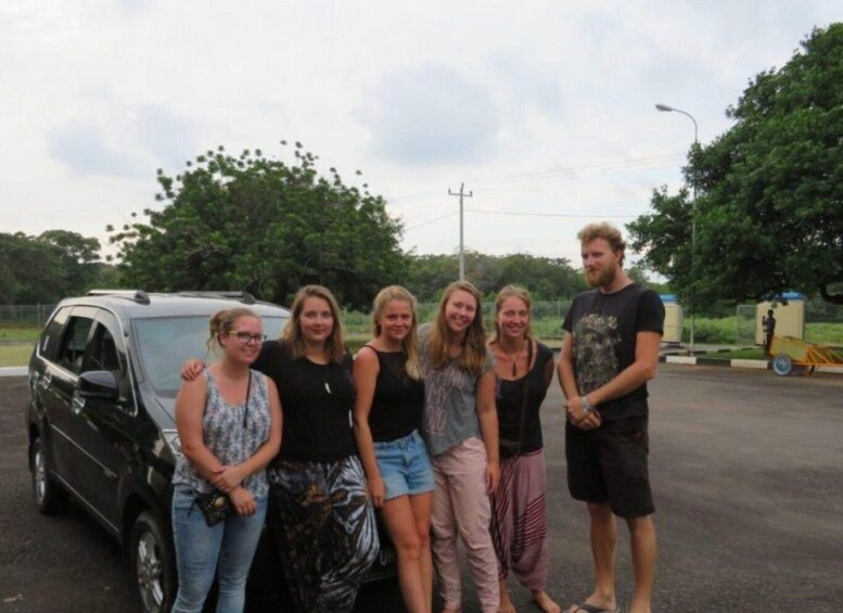 A group of people standing in front of a black car in an outdoor parking area with trees and a fence in the background. 3