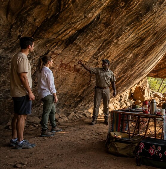 A cave with rock paintings, a guide pointing at them, and a table with camping gear. 1