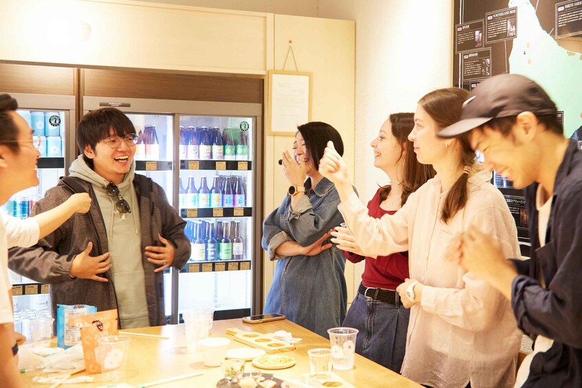 A group of people in a casual indoor setting with a refrigerator stocked with beverages and a table with food items. 3