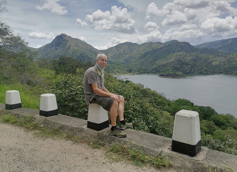 A man sitting on a concrete ledge with a lake and mountains in the background. 3