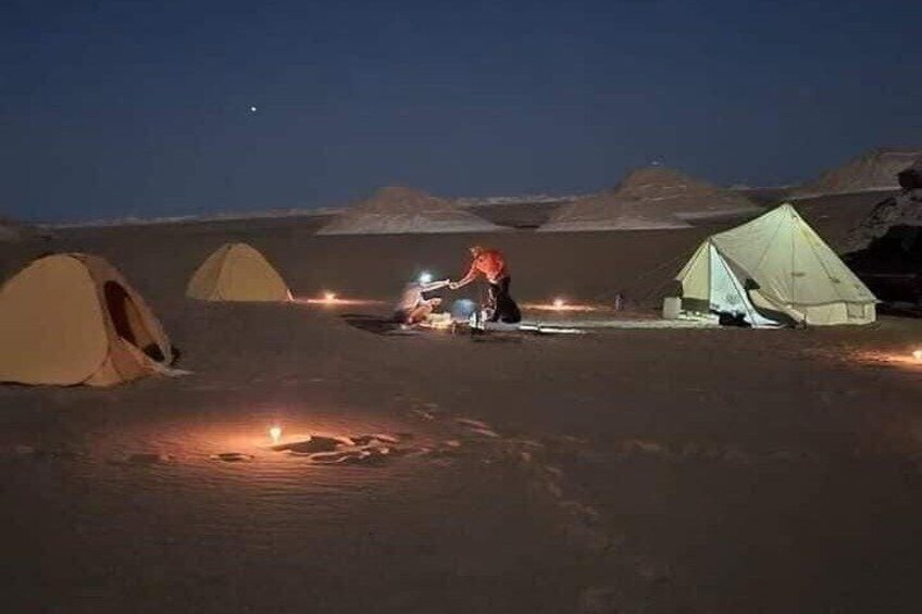 A desert campsite at night with tents and a person tending to a campfire. 1