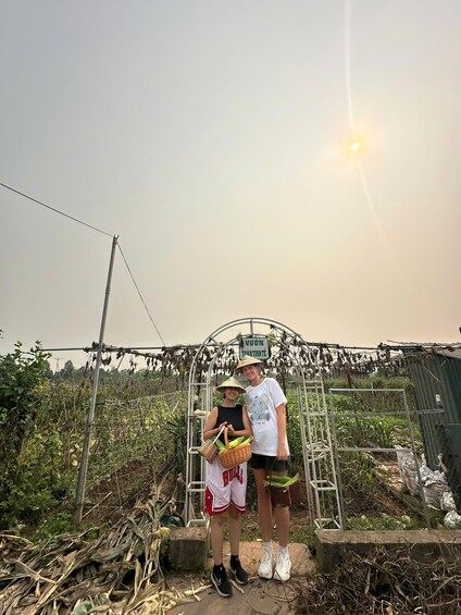 Two people standing in a field with a metal structure overhead and a sign that reads 'VUON TRANG TIENHIE.' 1