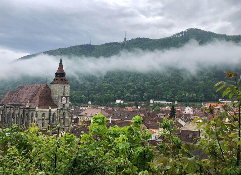 A historic church with a clock tower, surrounded by a town with red-roofed buildings, set against a backdrop of mist-covered hills. 1