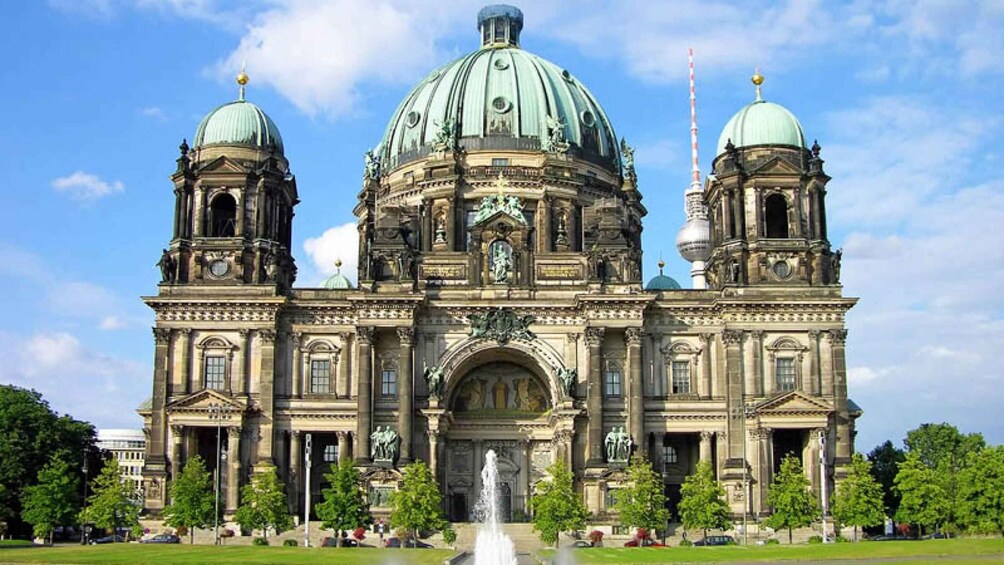 A grand historic building with two domes, a central archway, and a fountain in front. 2