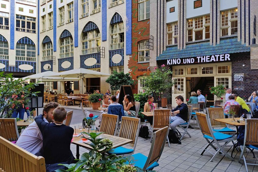 An outdoor cafe with tables and chairs, a building with 'KINO UND THEATER' signage, and people dining. 5