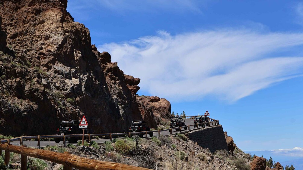 A winding mountain road with guardrails, a sharp bend warning sign, and a distant view of a rocky cliff. 3