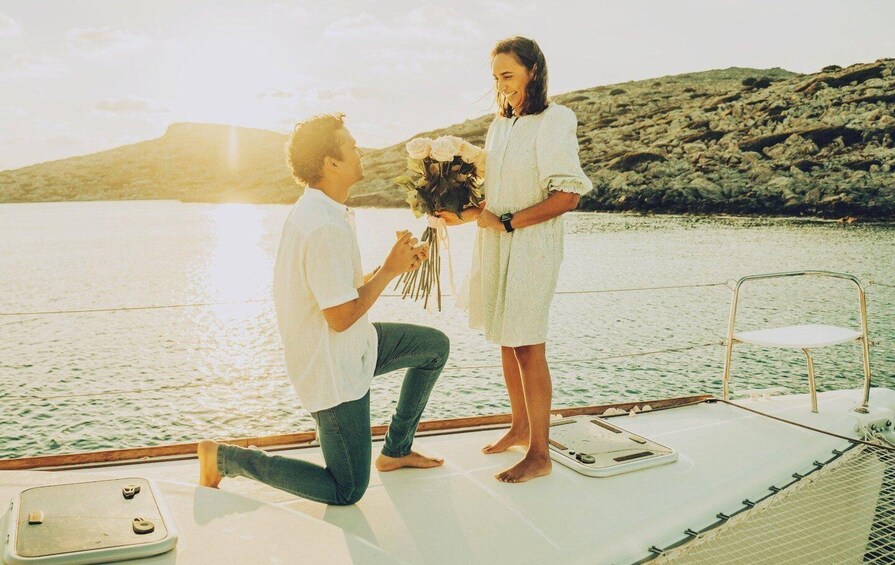 A person kneeling on a boat holding a bouquet, another person standing with a bouquet, and a rocky landscape in the background. 3