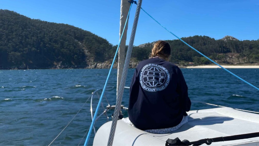 A person on a sailboat with a logo on the back of their jacket, surrounded by calm water and a forested coastline. 2