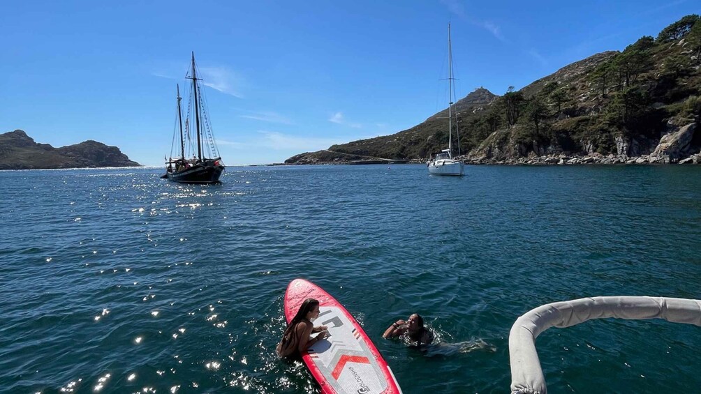 A coastal scene with a sailboat, a person on a surfboard, and another person swimming. 1