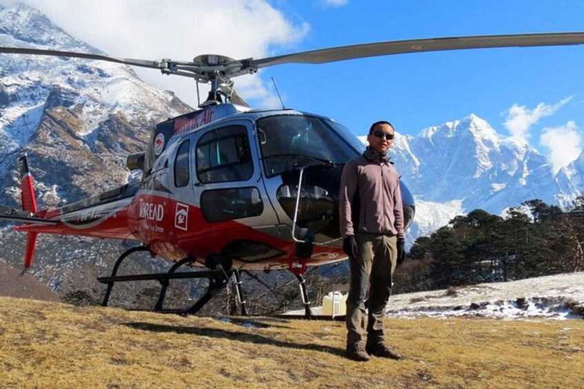A person standing in front of a helicopter with a mountainous landscape in the background. 1
