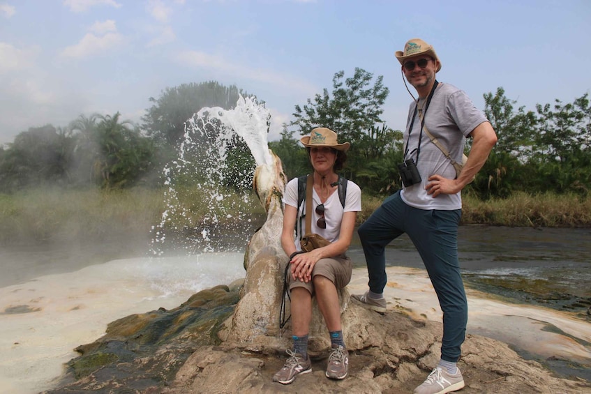 Zwei Personen sitzen auf einem Felsen neben einem Geysir, aus dem Wasserfontänen schießen. 2