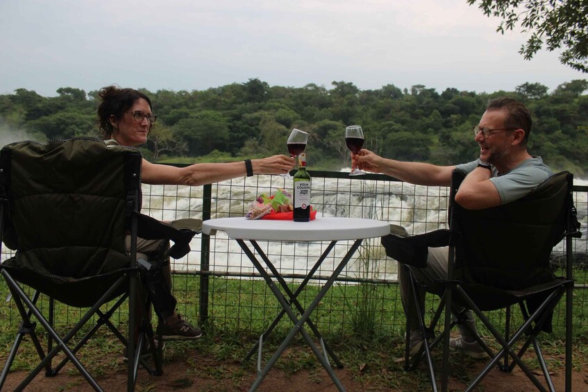 Zwei Personen stoßen mit Weingläsern an einem Tisch an, auf dem eine Flasche Four Cousins Sweet Red Wein steht, draußen mit einem Fluss und Grün im Hintergrund. 1