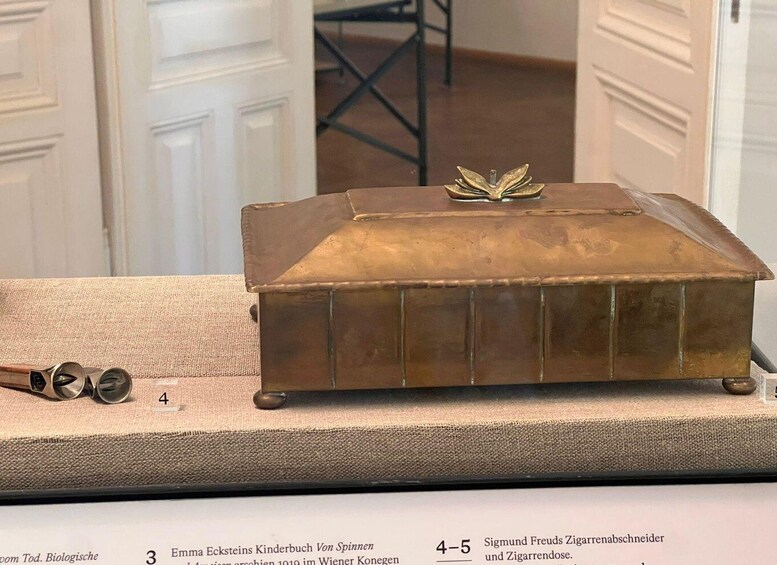 A brass box with a book on top, labeled '4-5 Sigmund Freud's Zigarettenabschnitt und Zigarrrendose.' 2