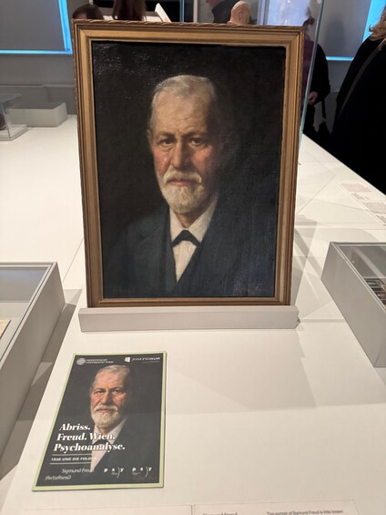 A framed portrait of a man in a suit, with a book titled 'Abriss. Freud. Wien. Psychoanalyse.' on a table. 1
