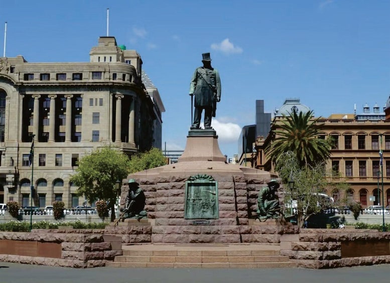 A statue of a man on a pedestal with a plaque, surrounded by stone steps and greenery, with buildings in the background. 3