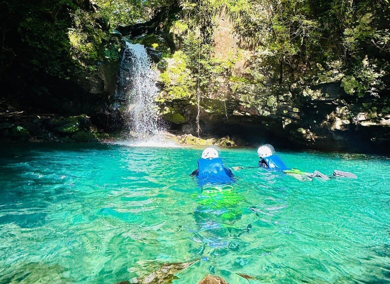 Two people swimming in clear turquoise water with a waterfall in the background. 1