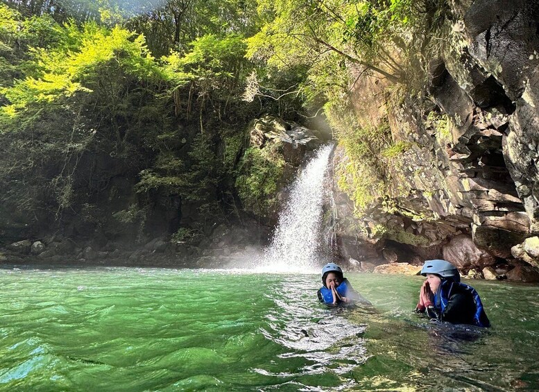 Two people in water near a waterfall surrounded by greenery. 4