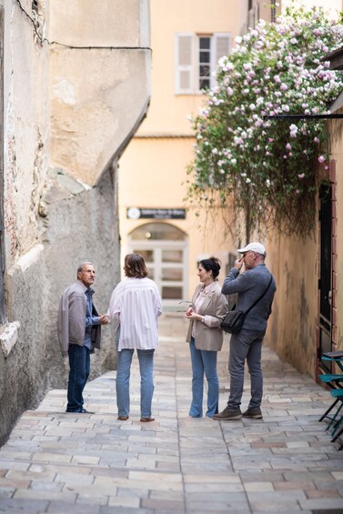 A narrow alley with people conversing, a building with a sign, and flowering vines. 4