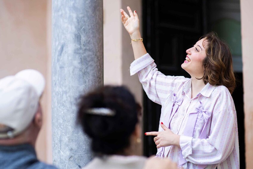 A person gesturing upwards, wearing a striped shirt with a bow, standing in front of a building with a column. 5