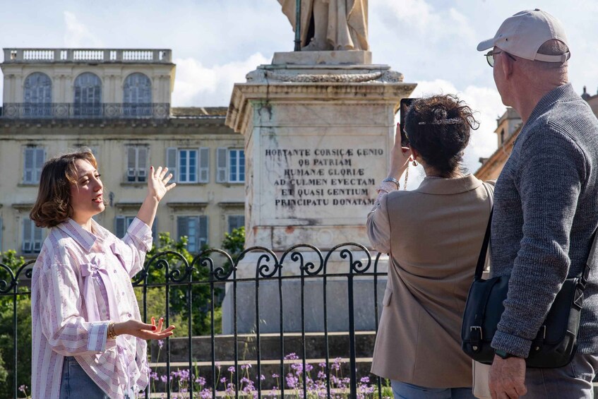A group of people standing in front of a statue with an inscription, a building with arched windows, and a fence with purple flowers. 3