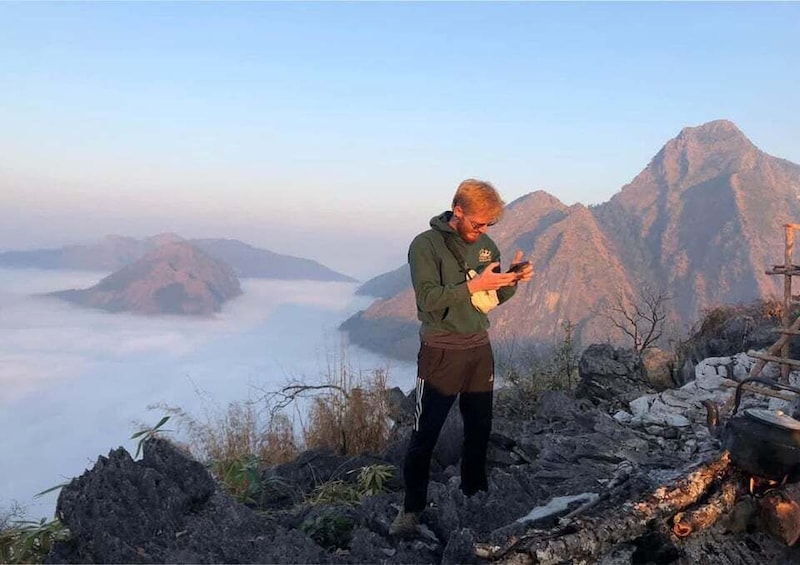 A person standing on a rocky outcrop, using a smartphone, with a mountainous landscape and clouds in the background. 3
