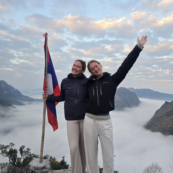 Two people standing on a mountain with a flag, clouds below, and a valley. 1