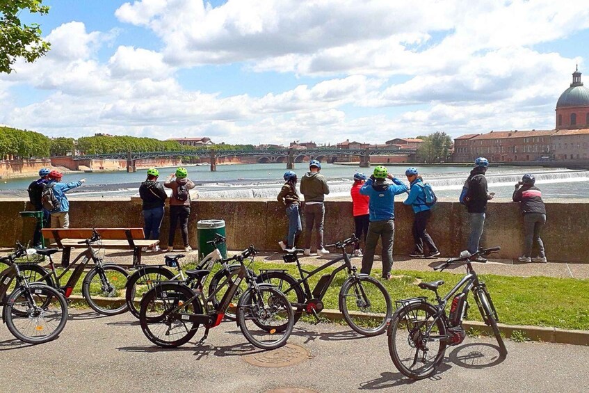 A group of cyclists and pedestrians by a riverside with bicycles parked and a bridge in the background. 1