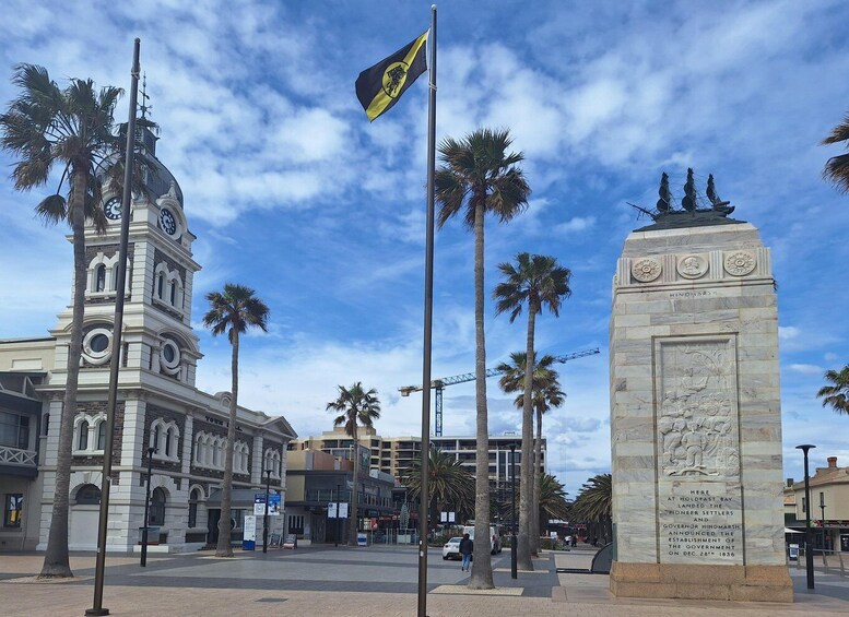 A historic building with a clock tower, palm trees, and a monument with inscriptions. 1
