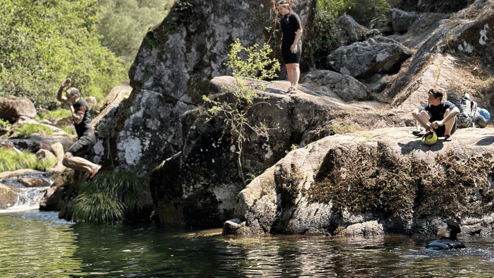People near a rocky riverbank, some standing and others sitting, with a flowing river in the foreground. 5