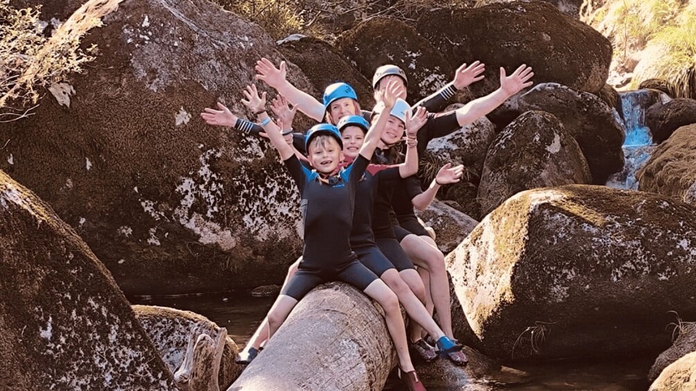 A group of people sitting on a large rock with their arms raised, surrounded by other rocks and a small waterfall. 1