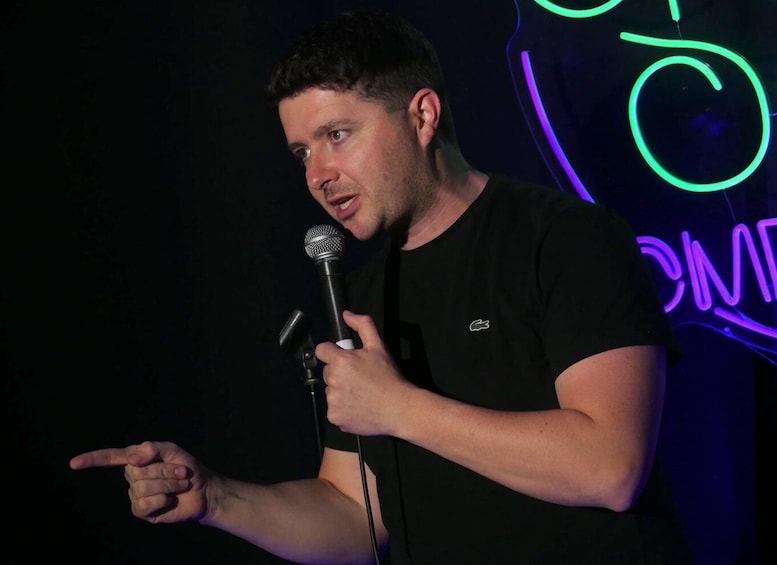 A person holding a microphone, wearing a black t-shirt with a small logo, in a dimly lit environment with neon signs in the background. 2
