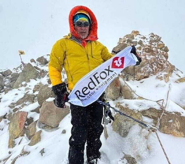 A person in a yellow jacket holding a RedFox banner on a snowy mountain peak. 4