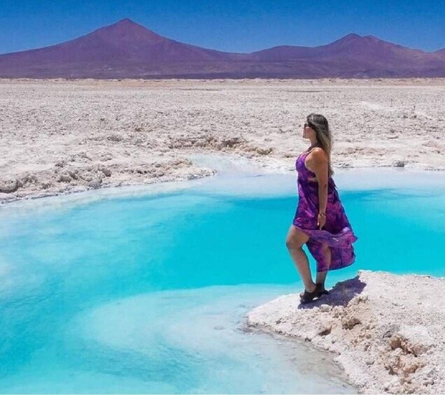 A person standing on a rocky outcrop by a turquoise pool with mountains in the background. 5