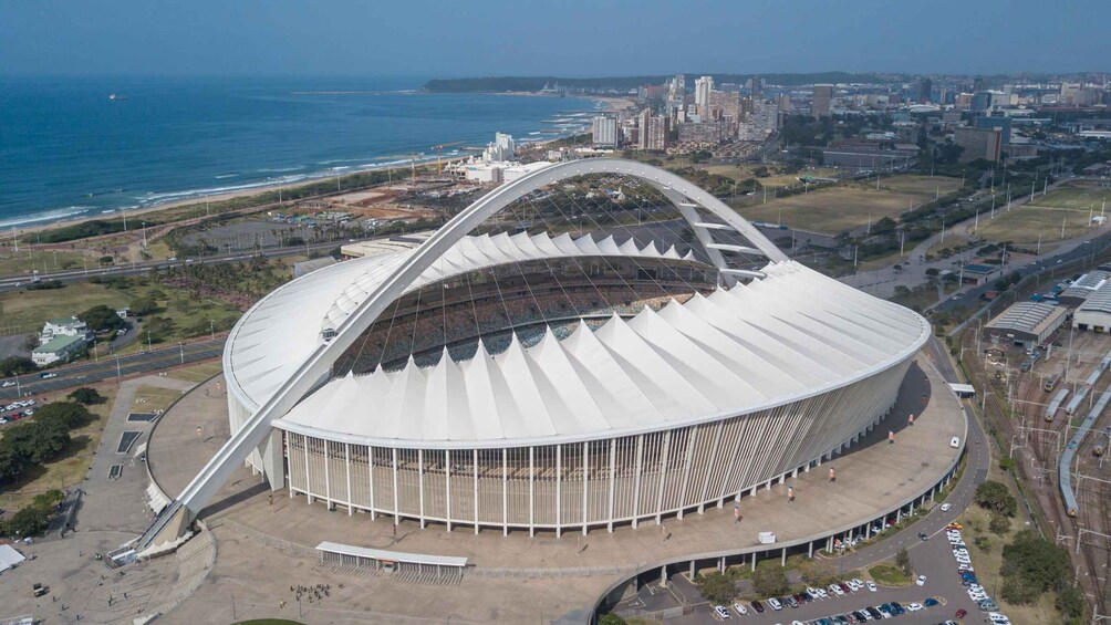A large stadium with a distinctive arch design, surrounded by roads and a coastal cityscape. 2