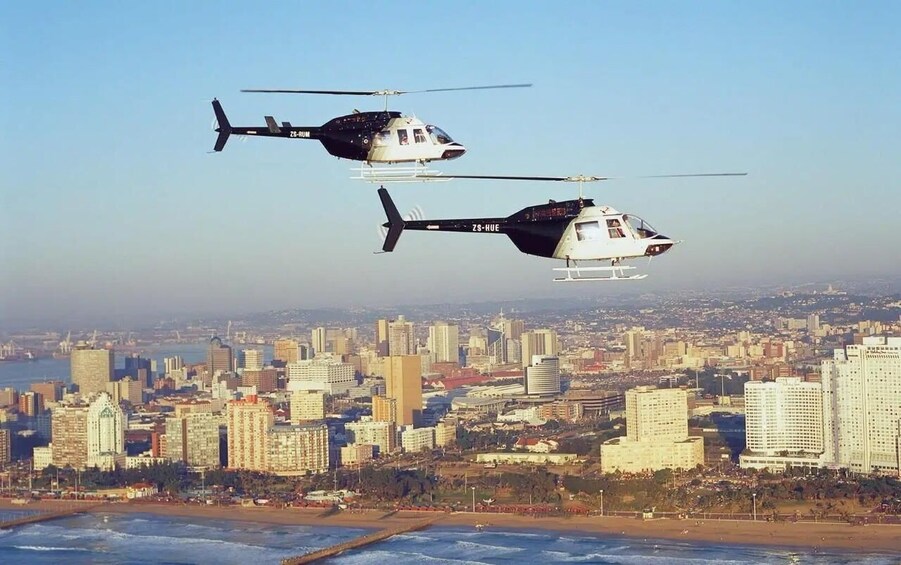 Two helicopters flying over a city with a beach and ocean in the foreground. 1