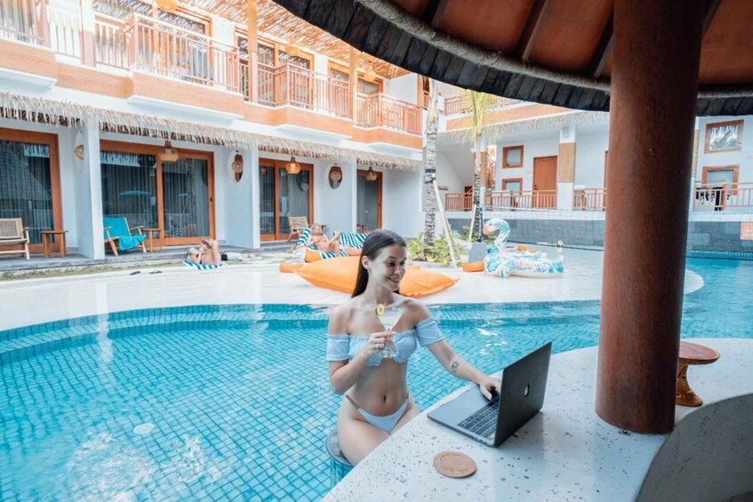 A person sitting by a poolside with a laptop, surrounded by a resort with multiple balconies and lounge chairs. 1