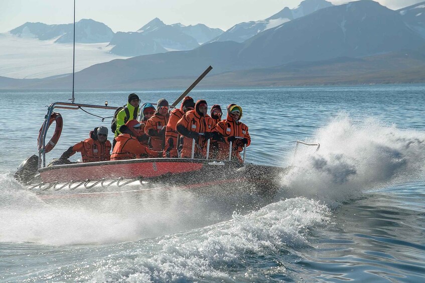 A group of people in orange life jackets on a small boat moving through water with mountains in the background. 4