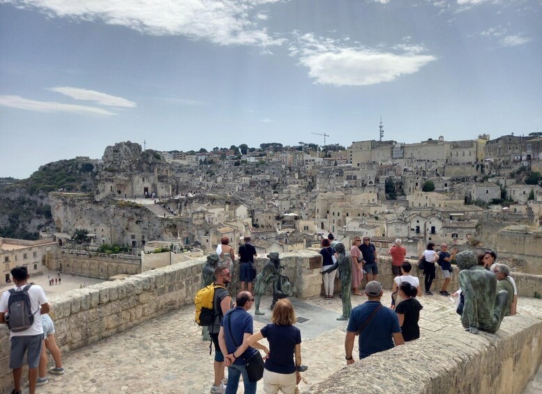 A group of tourists on a stone platform overlooking a historic town with layered buildings and a prominent rock formation. 3