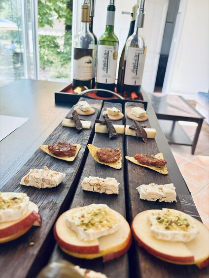 A wooden table with various cheese and meat appetizers, and bottles of wine in the background. 4